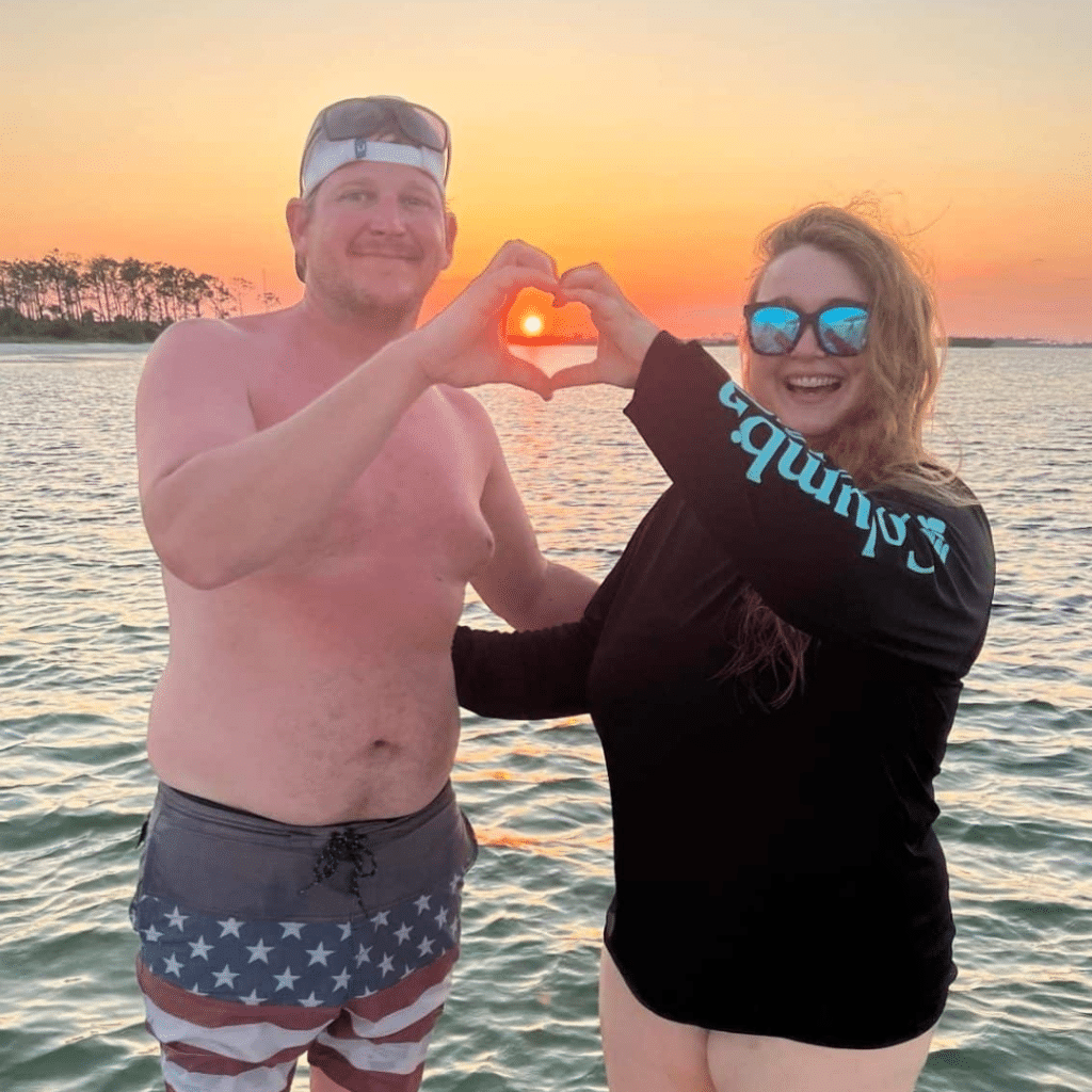Couple enjoying a Sunset Dolphin Tour & Cruise in Panama City Beach, forming a heart shape with their hands around the setting sun over the water.