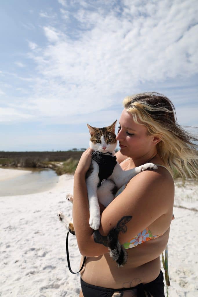 Woman holding a cat in a harness on a white sandy beach under a blue sky, enjoying one of the unexpected but pet-friendly moments while exploring dog friendly things to do in Panama City Beach.