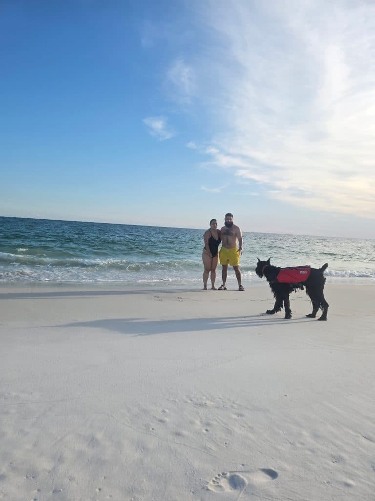 A couple stands along the shoreline at Shell Island while their dog in a red life vest explores the white sand, showcasing one of the top dog friendly things to do in Panama City Beach on a Flippin' Awesome Adventures tour.